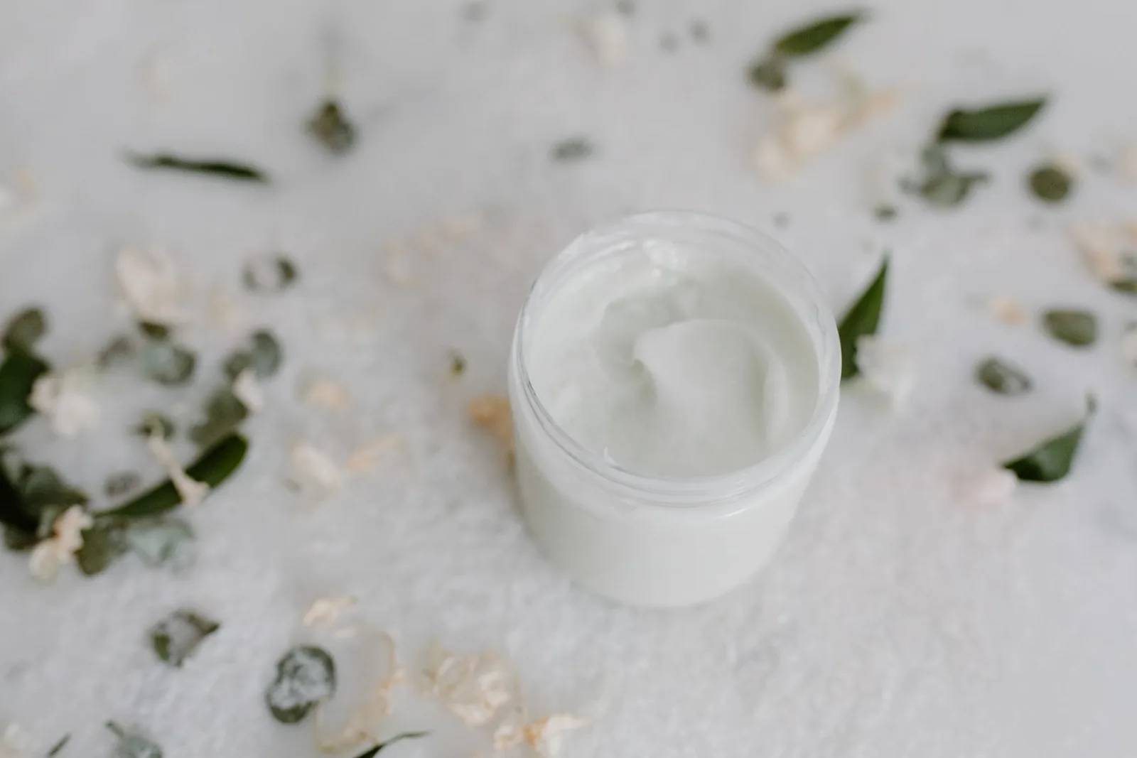 Open white cream jar surrounded by scattered green leaves