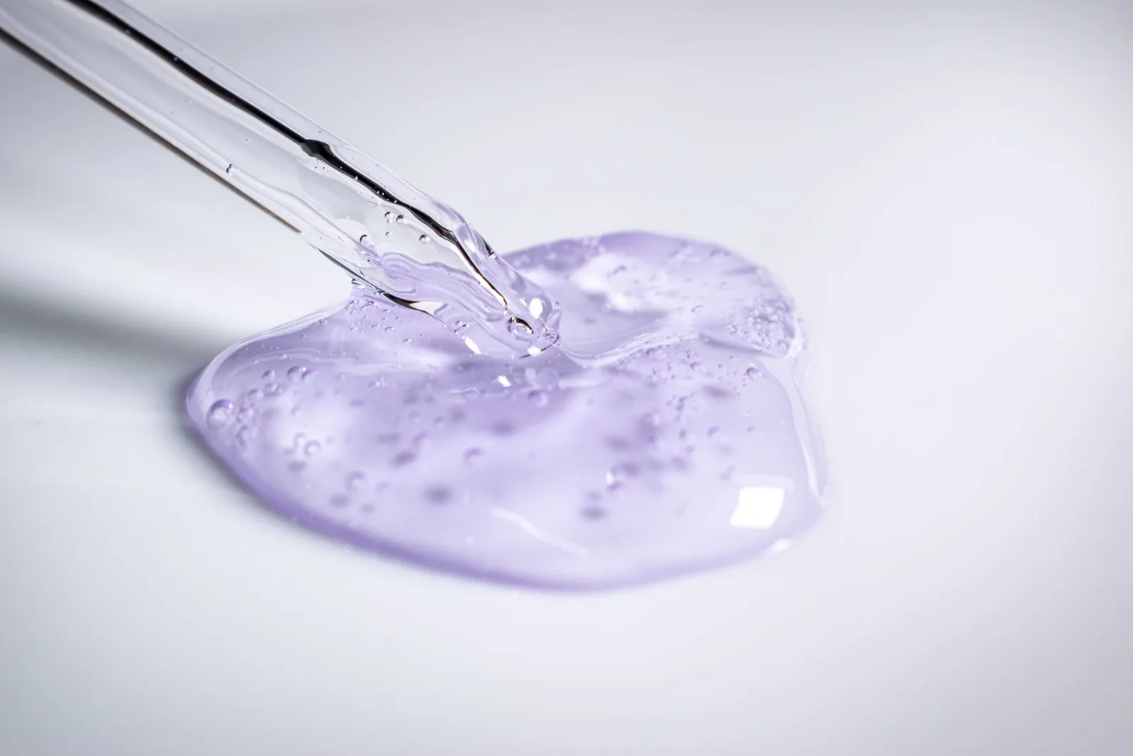 Pipette dispensing clear lilac cosmetic gel onto a white surface